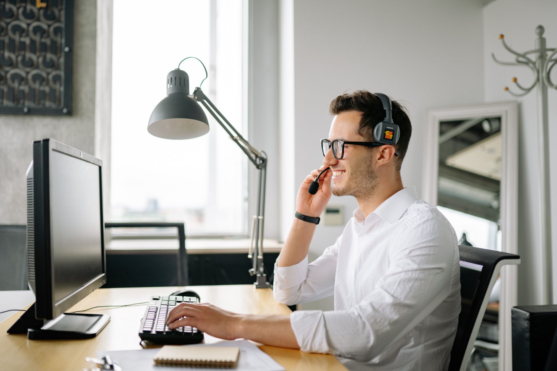 Lächelnder Mann mit Headset arbeitet in einem hellen Büro am Computer.